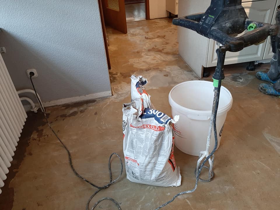 Materialien und Werkzeuge für ein Hausrenovierungsprojekt, darunter Bodenverlegungsmaterialien, ein teilweise verbrauchter Beutel mit Spachtelmasse und ein Mixer, liegen auf dem Boden verstreut. T&K WOHNSTDIO - Raumausstattung, Bodenbeläge, Vinylboden, Parkett, Gardinen - Saarburger Straße 25, 67071 Ludwigshafen