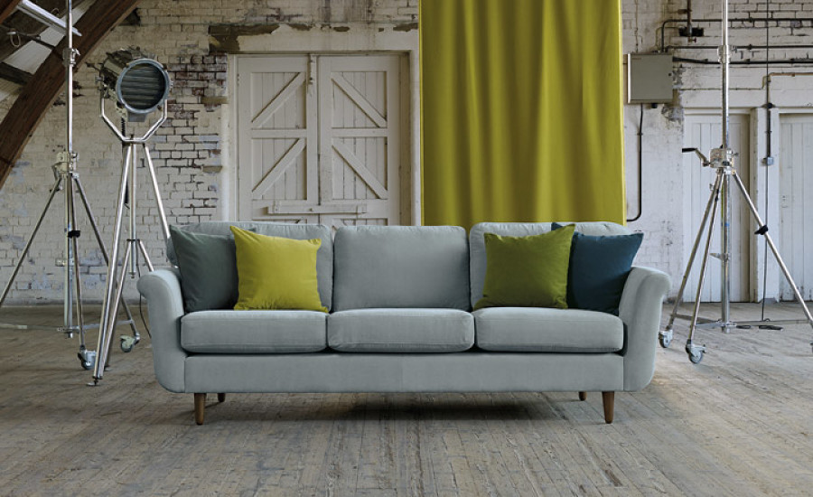 Bild Aufmaß Ein modernes Wohnzimmer-Set mit einer grauen Couch mit gelben und grünen Kissen und Studiobeleuchtung vor einer strukturierten weißen Ziegelwand mit drapiertem gelben Vorhang. T&K WOHNSTDIO - Raumausstattung, Bodenbeläge, Vinylboden, Parkett, Gardinen - Saarburger Straße 25, 67071 Ludwigshafen