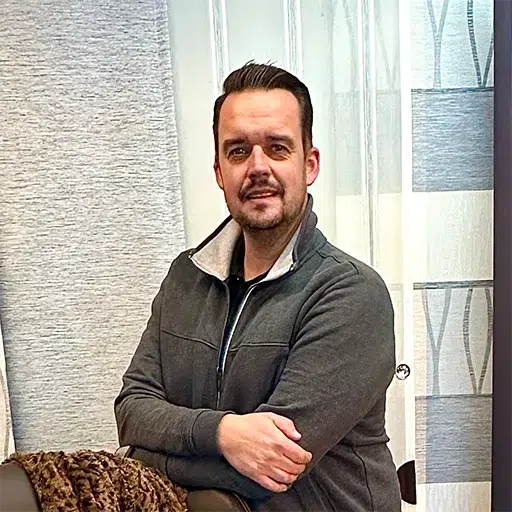 Ein Mann mit kurzen Haaren und einer dunklen Jacke sitzt mit verschränkten Armen vor gemusterten Vorhängen und verkörpert durch sein gelassenes Auftreten die Essenz von „Über Uns“. - T&K WOHNSTDIO - Raumausstattung, Bodenbeläge, Vinylboden, Parkett, Gardinen - Saarburger Straße 25, 67071 Ludwigshafen