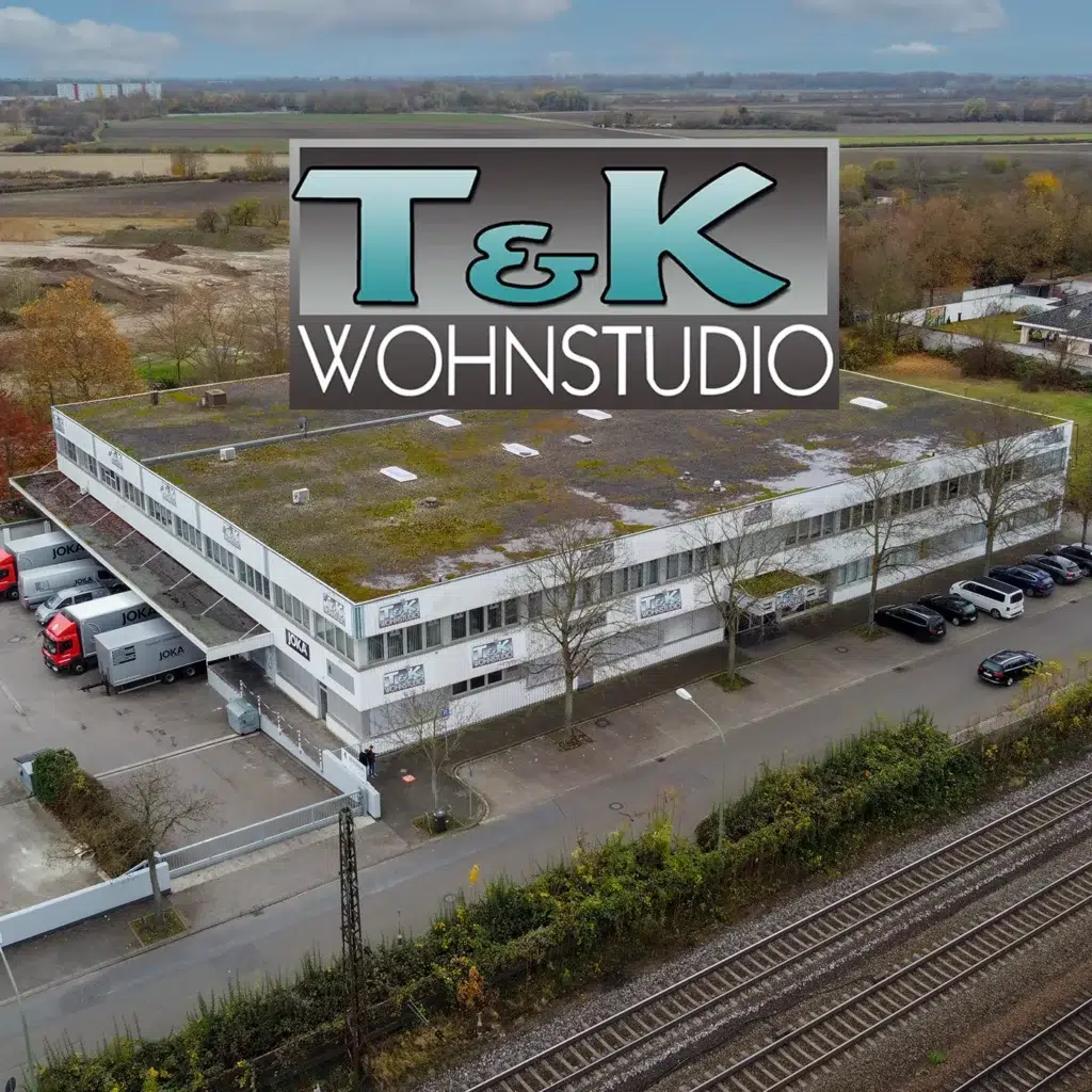 Aus der Luft ist ein rechteckiges Gebäude mit einem markanten „T&K Wohnstudio“-Schild auf dem Dach zu erkennen. In der Nähe parken mehrere Fahrzeuge, eingebettet zwischen Bahngleisen und Feldern, was eine Atmosphäre hervorruft, die der Mischung aus Zweckmäßigkeit und malerischer Umgebung von Schads Wohnträume ähnelt. - T&K WOHNSTDIO - Raumausstattung, Bodenbeläge, Vinylboden, Parkett, Gardinen - Saarburger Straße 25, 67071 Ludwigshafen